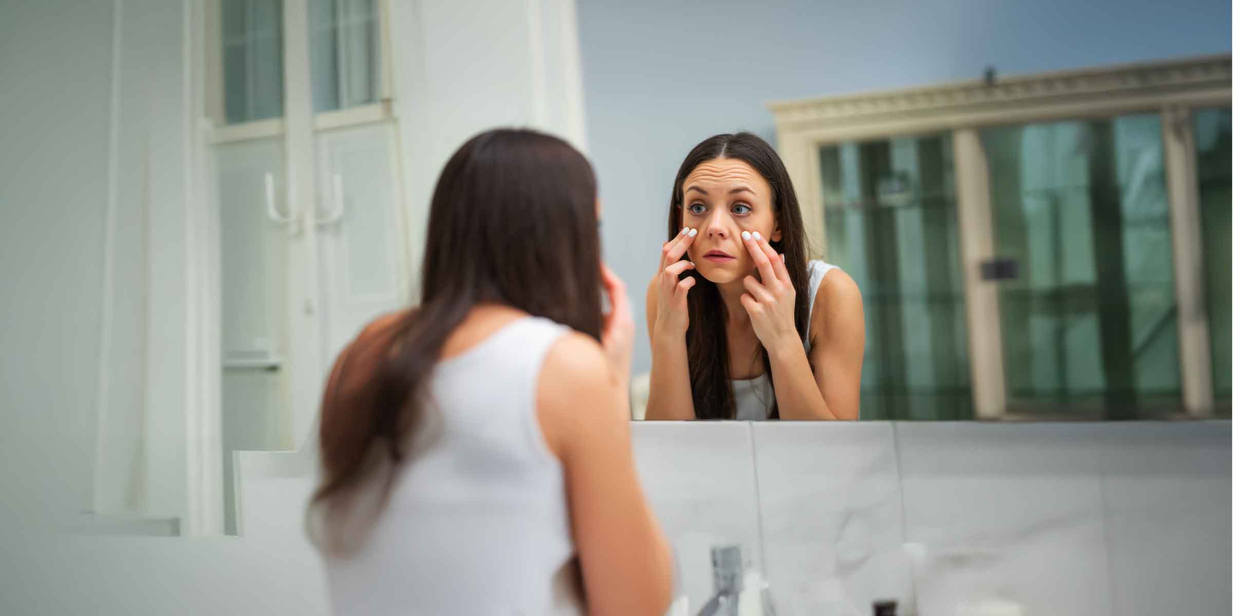 Woman checking face in mirror for under-eye puffiness, a common sign of kidney fluid imbalance.