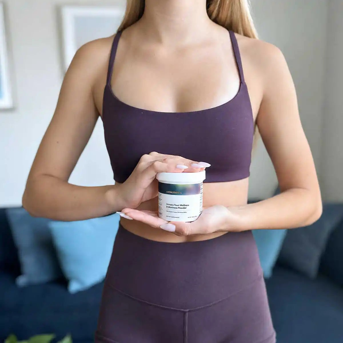 A woman in a dark purple sports bra and matching leggings holds a white jar of Urinary Tract Wellness D-Mannose Powder