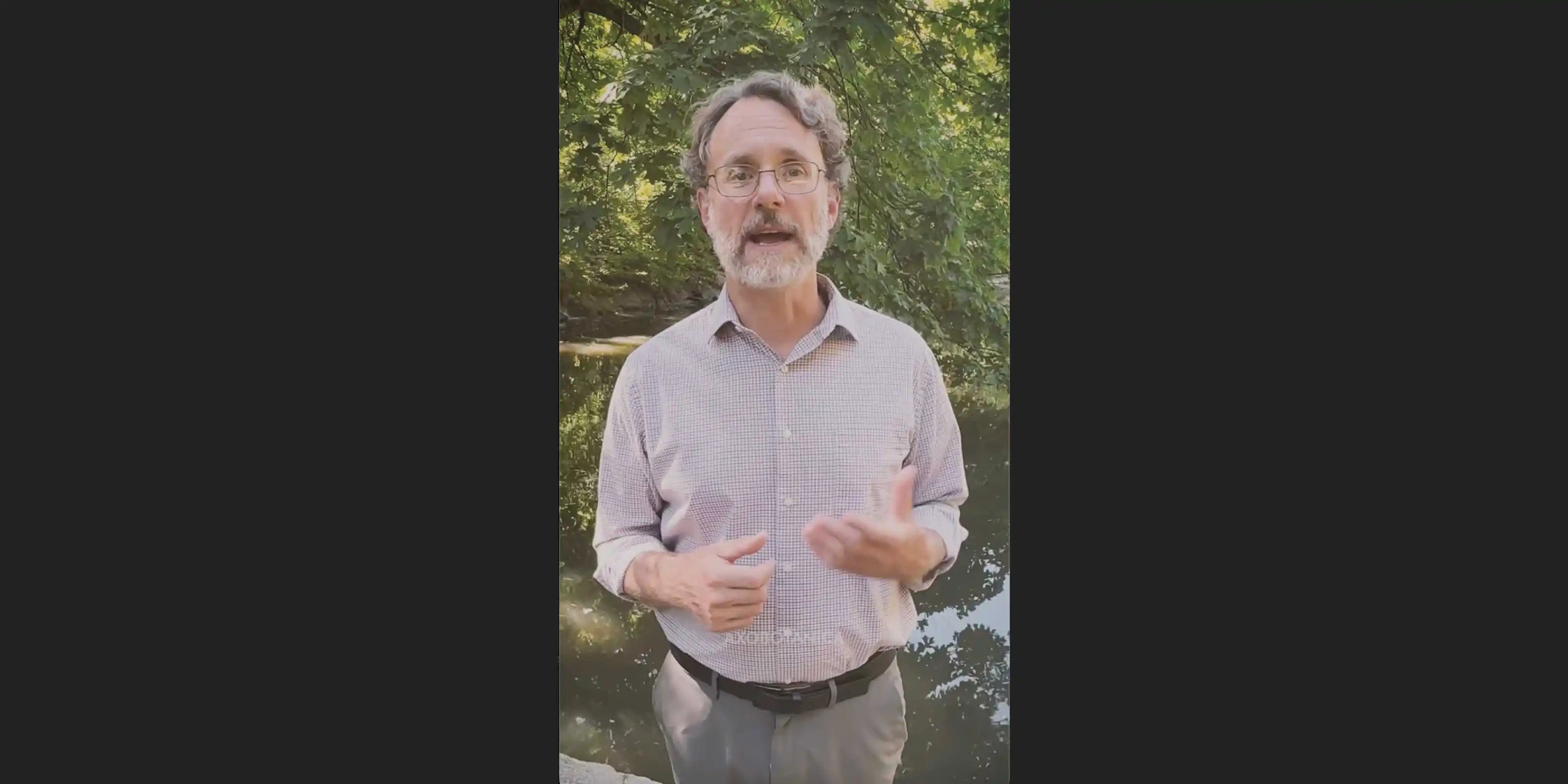 A man with glasses, a gray beard, and a light-colored button-down shirt stands outdoors in front of green trees and water, gesturing with his hands.
